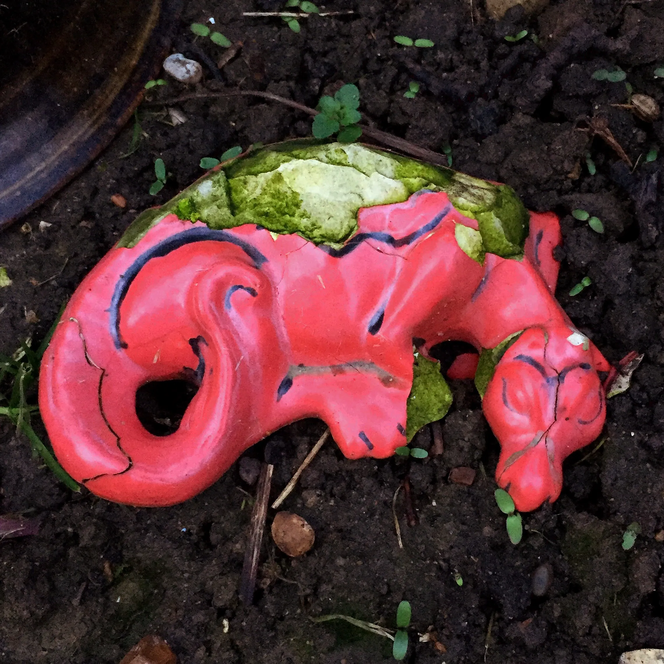 A weathered decorative object painted deep red with dark markings, curled like an ornament, set against a moody dark background with hints of moss green