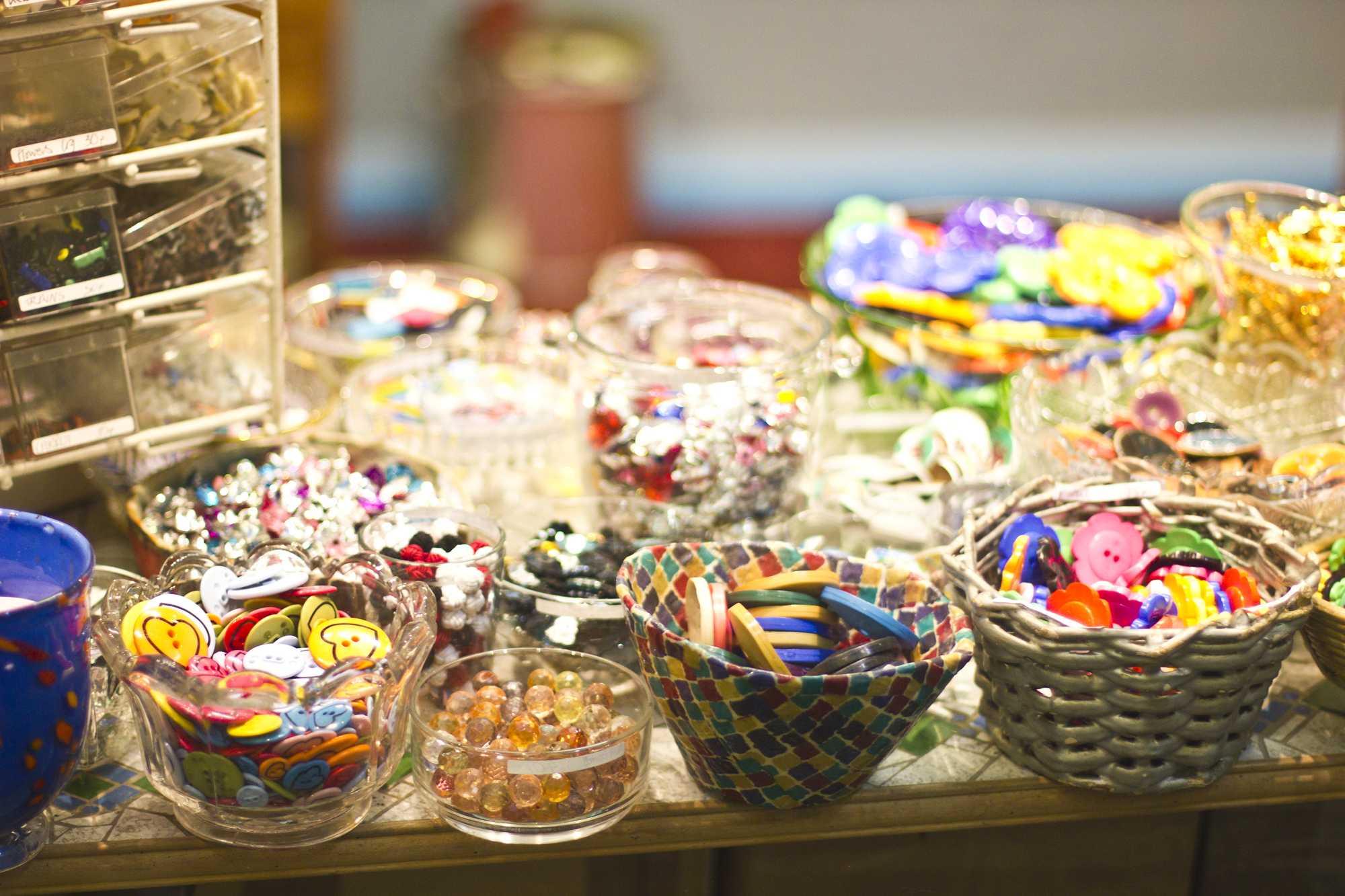 A colorful assortment of buttons, beads, and craft supplies gathered in glass bowls and woven baskets on a wooden table